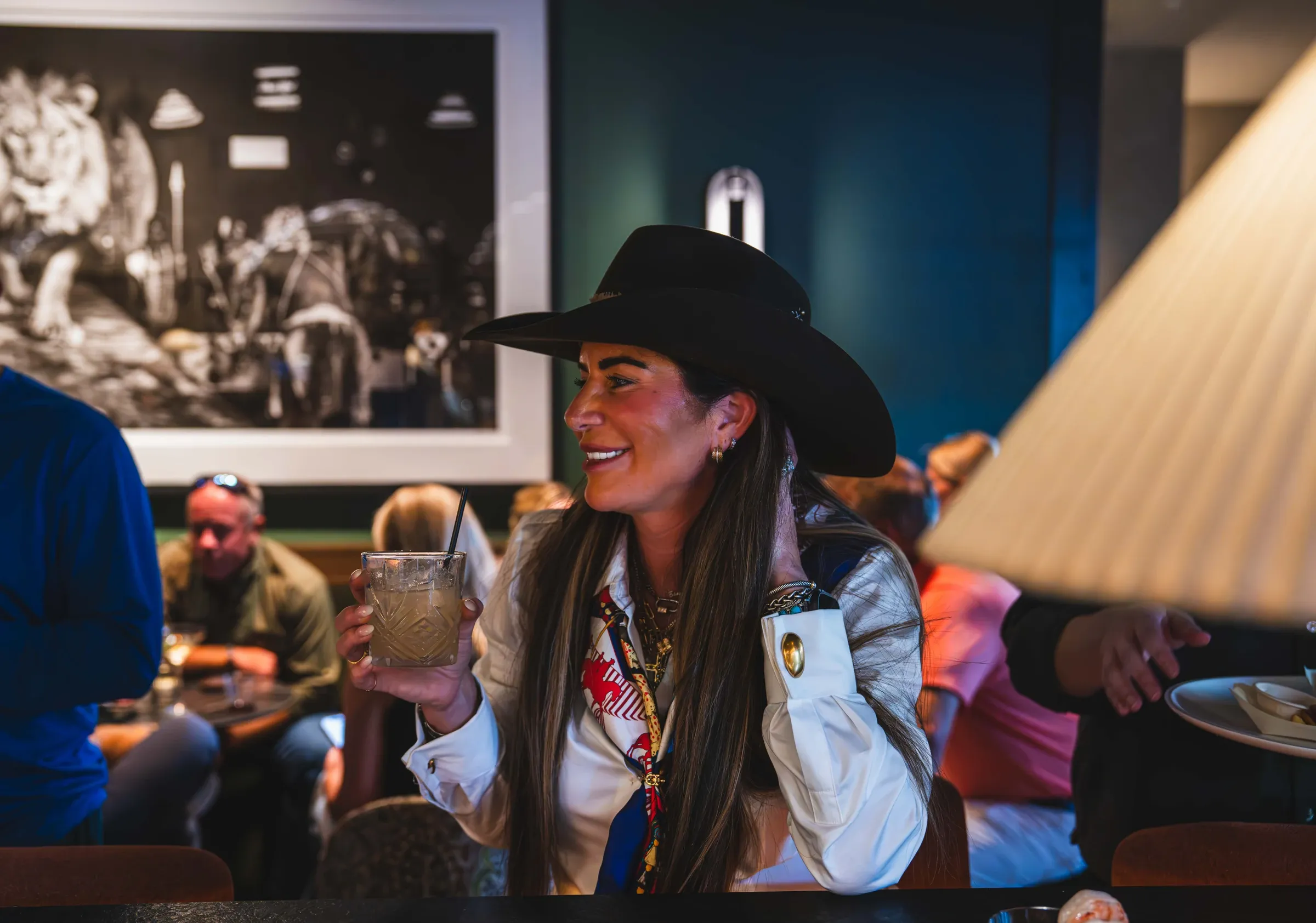 A guest in a black cowboy hat smiles over a cocktail at the bar, the lion print on the wall behind