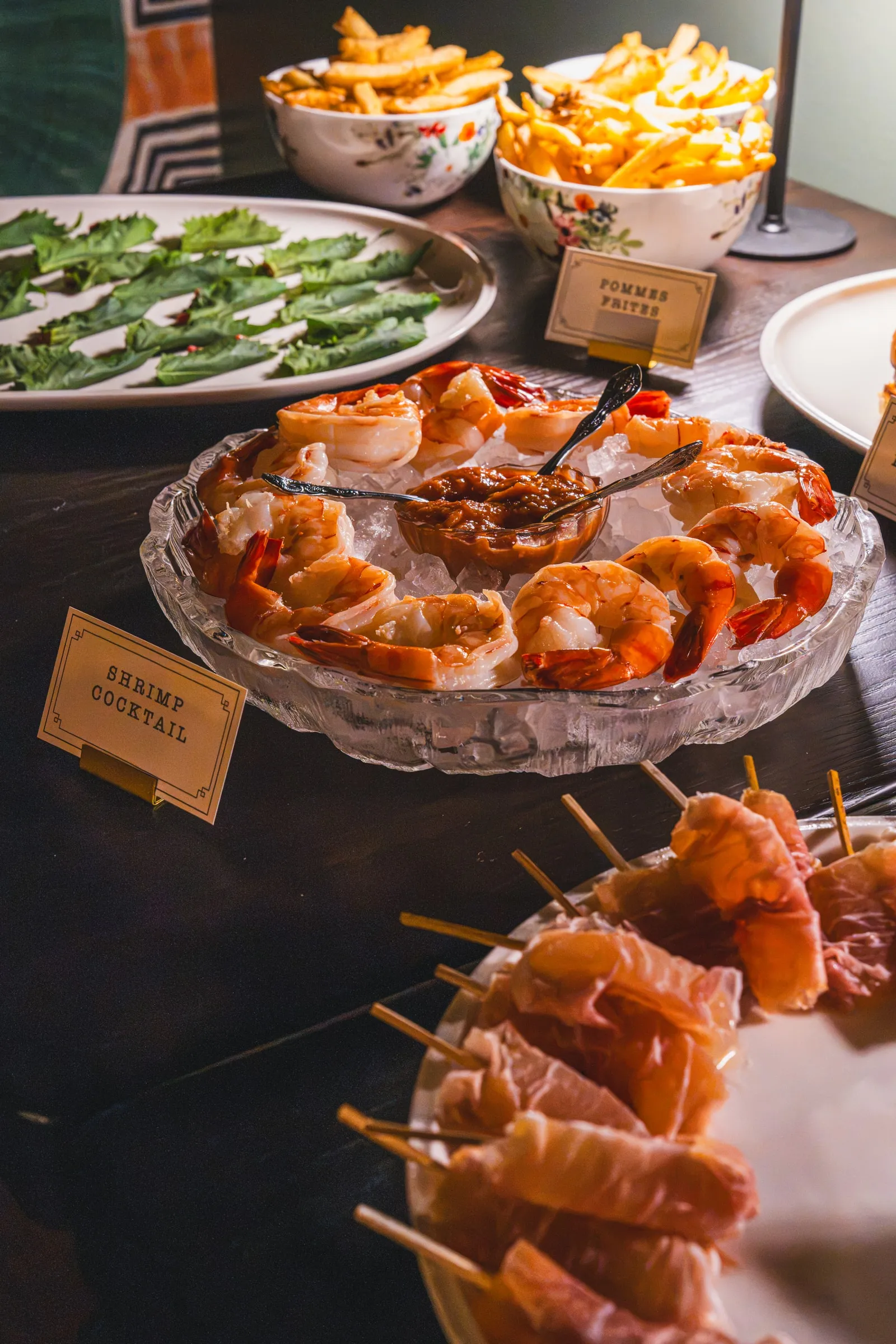 An event spread of shrimp cocktail on ice, prosciutto skewers, fries, and snap peas