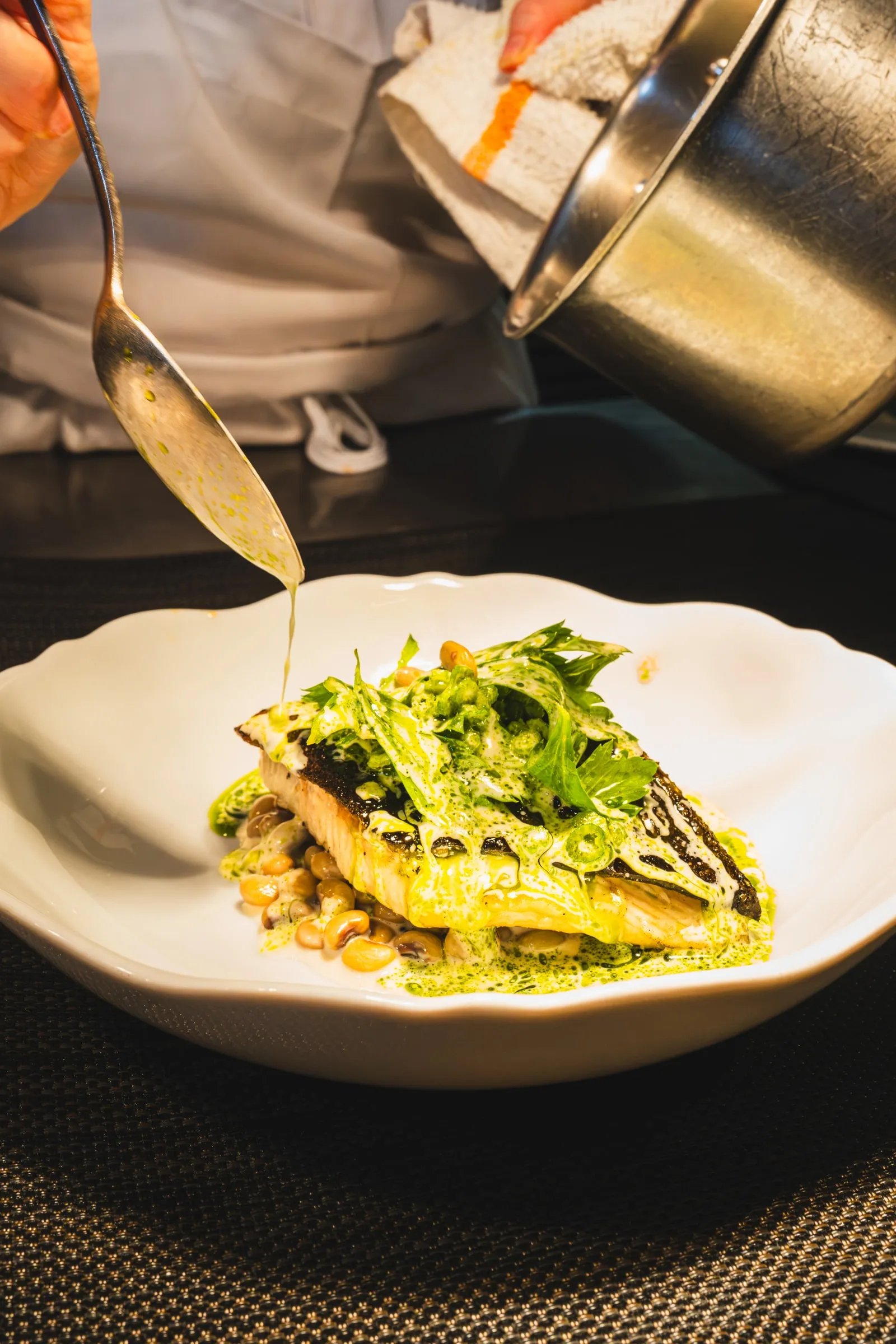 A cook spoons sauce over halibut with greens in a white bowl